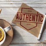 A cup of tea with a lemon in it, sitting on a table next to a small sign saying “Authentic.”