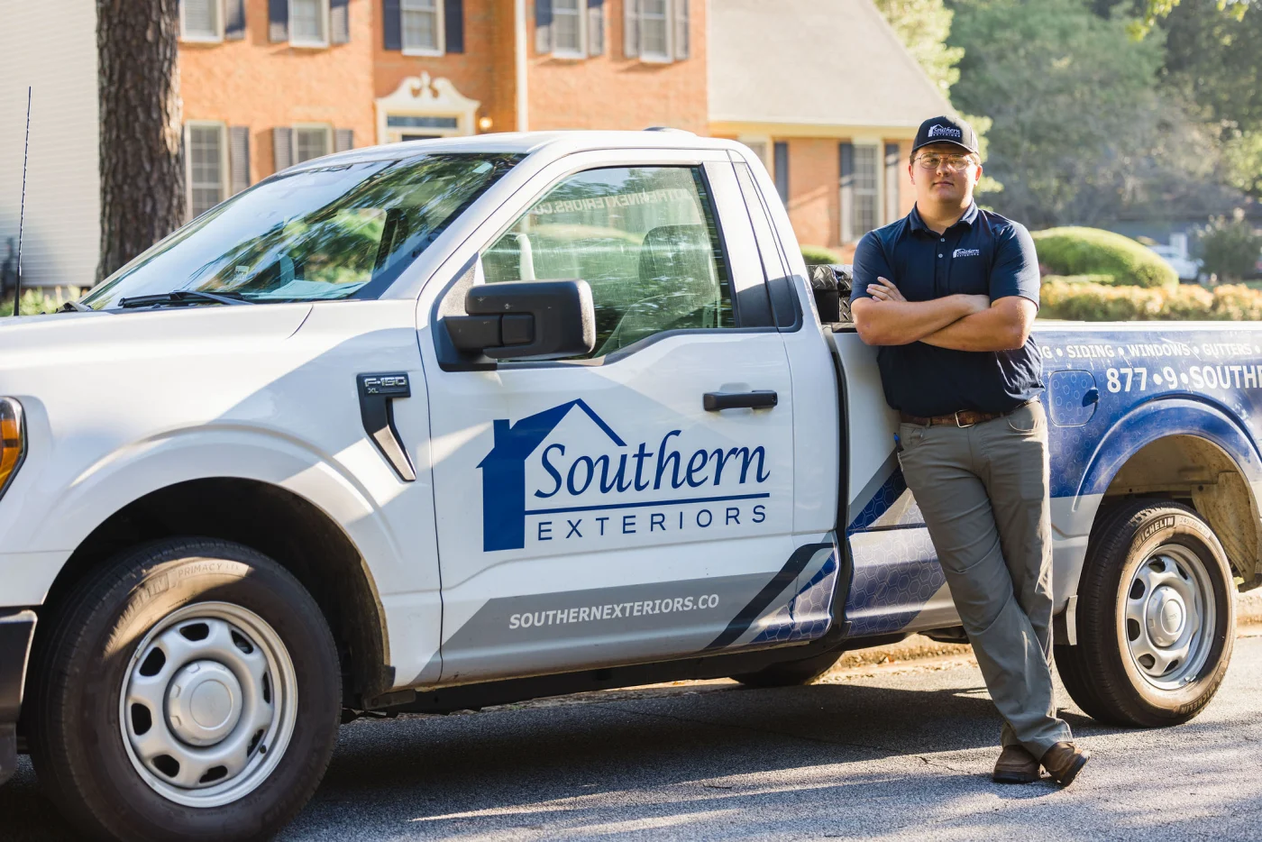 Amazing Remodeling Contractor | Columbia, SC | Southern Exteriors A team member wearing a branded company shirt and cap leans against a white Southern Exteriors pickup truck on a bright residential street, highlighting the dependable service of an amazing exterior remodeling contractor.