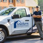 A uniformed professional stands with crossed arms while leaning against a branded Southern Exteriors pickup truck in a residential neighborhood, representing the dedicated service of a local exterior remodeling contractor.