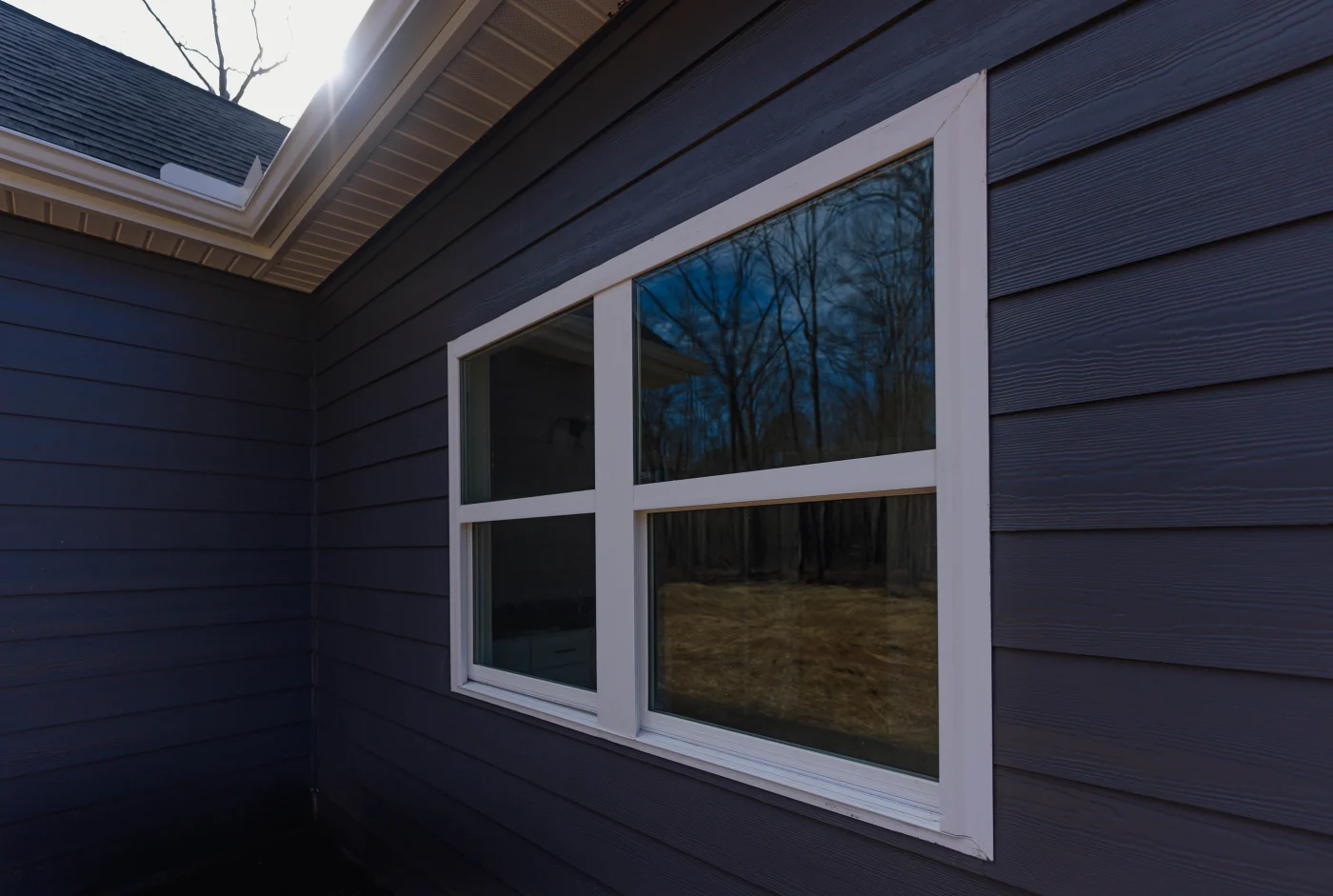 Elegant Window Replacements | Columbia, SC | Southern Exteriors A side-angle view captures a clean white-trimmed double-pane window set against dark horizontal siding, beautifully reflecting the surrounding trees and bright sky.