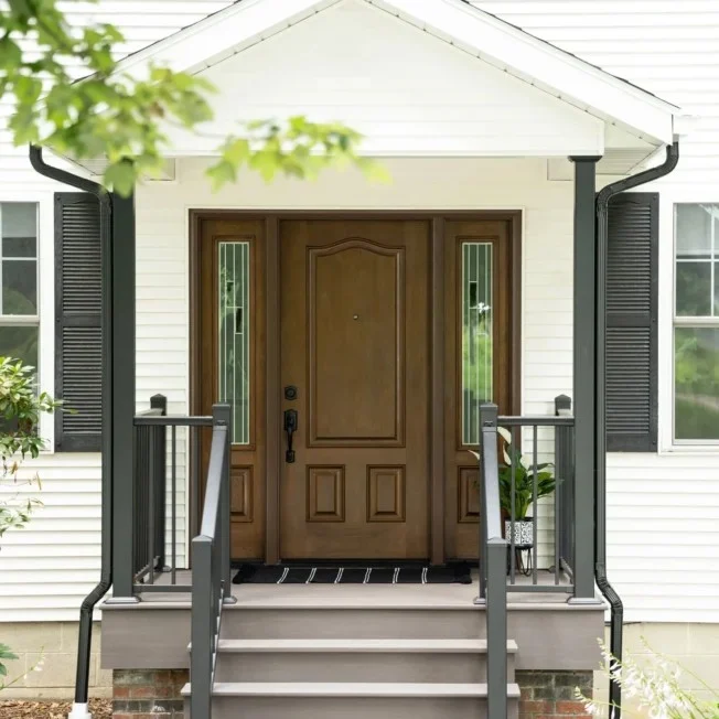 Exceptional New Construction Services | Columbia, SC | Southern Exteriors A handsome wood entry door with matching glass sidelights serves as the focal point for this welcoming front porch, beautifully framed by white horizontal siding, a gabled roof overhang, and dark railings and shutters.