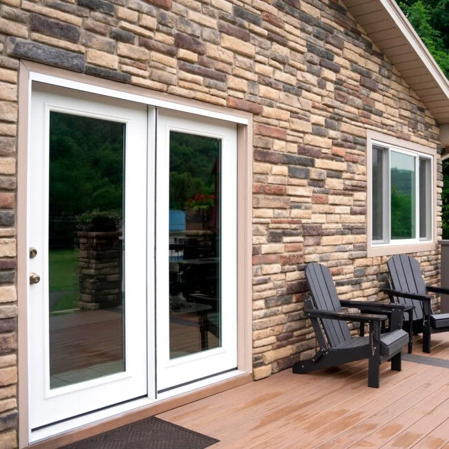 Stunning Door Replacements | Columbia, SC | Southern Exteriors Elegant white double patio doors with large glass panes open up to a wooden deck alongside a beautiful stacked stone accent wall featuring two dark Adirondack chairs.