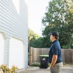 A professional representing a local remodeling contractor stands in a driveway with his hands in his pockets while assessing the light gray siding above two white garage doors.