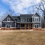 A wide shot showcases a beautiful two-story home transformation featuring dark charcoal vertical siding, white window trim, and a brick foundation skirt completed by an expert remodeling contractor.